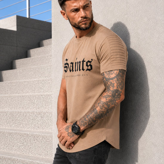 Male model wearing a beige SAINTS graphic T-shirt with black jeans in an urban casual outfit