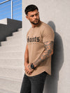 Male model wearing a beige SAINTS graphic T-shirt with black jeans in an urban casual outfit