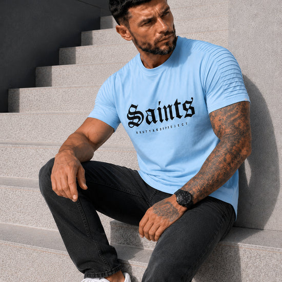 Male model sitting in a light blue SAINTS graphic T-shirt with black pants and white sneakers