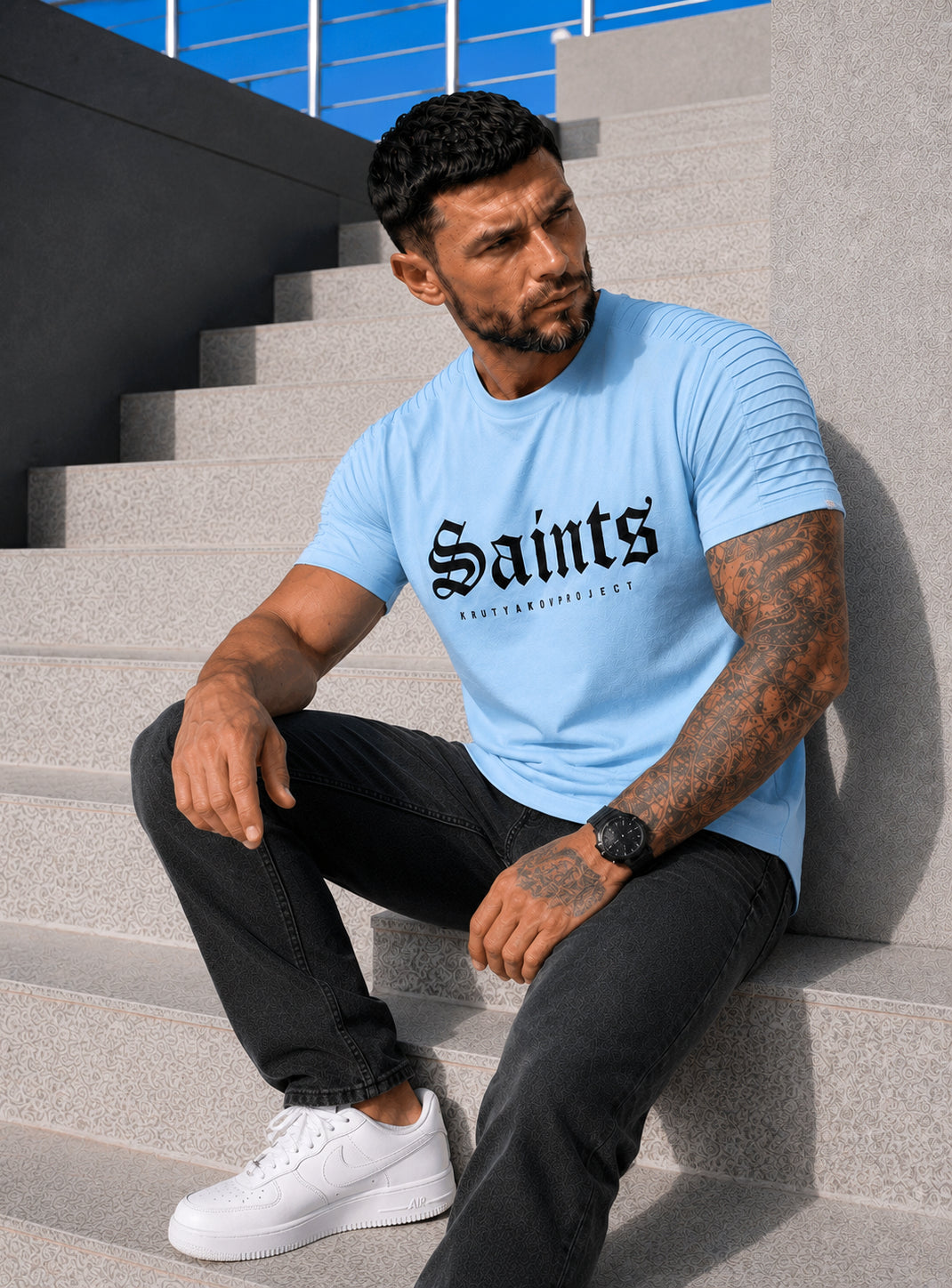 Male model sitting in a light blue SAINTS graphic T-shirt with black pants and white sneakers